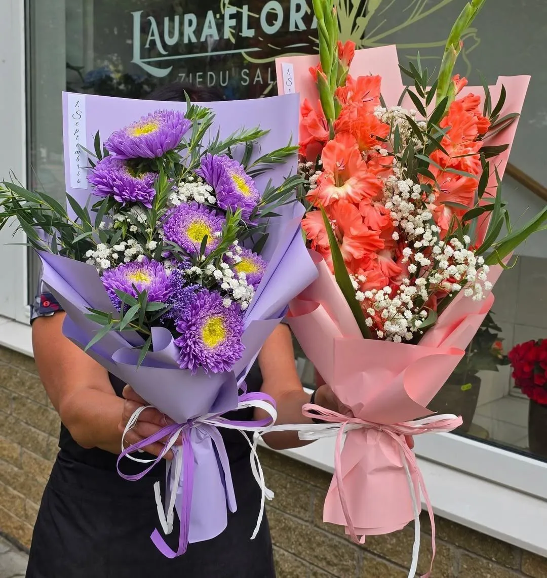 Divi ziedu pušķi – violetas asteres un oranžas gladiolas ar ģipsenēm – pie salona „LauraFlora” skatloga